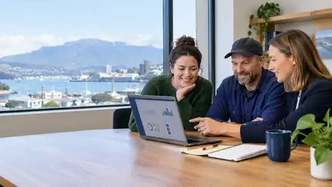 Three Tasmanian small-business colleagues review digital business performance charts together at a laptop in a Hobart office overlooking the harbour.